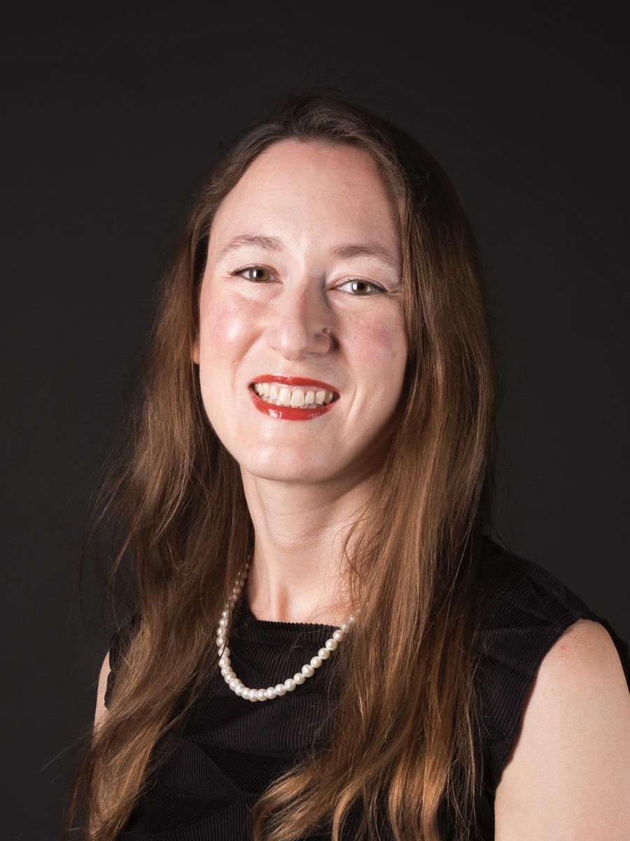 Porträt einer Frau mit langen braunen Haaren. Sie lächelt freundlich, trägt ein schwarzes Kleid und eine weiße Perlenkette. Der Hintergrund ist einheitlich dunkel, was den Fokus auf ihr Gesicht und den Oberkörper lenkt.